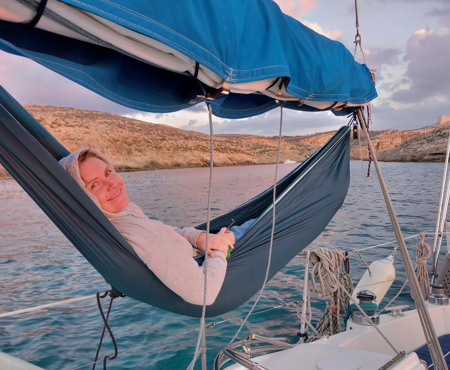 A guest in a hammock slung between the shrouds, with Gozo cliffs behind.